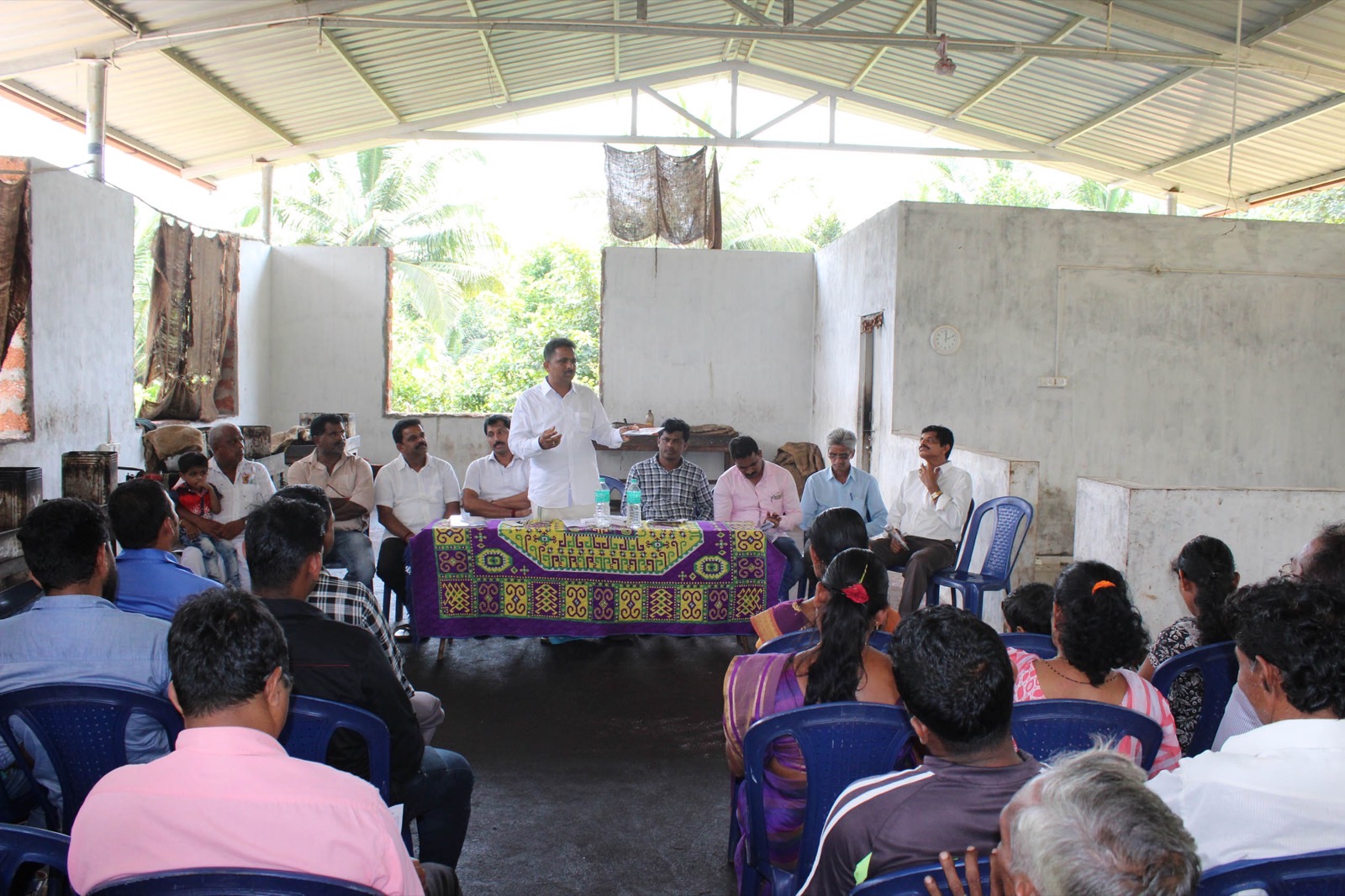 Udaya Shetty Muniyal addressing a seated village gathering under a tin roof